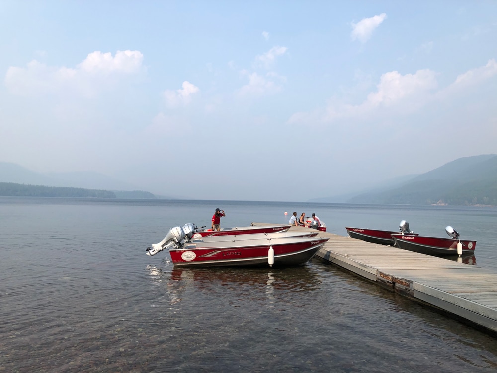 View of Lake McDonald, from the same spot, in Glacier National Park in August 2021. THe sky is hazy, fewer boats are along the dock and three people are gathered at the end of the dock.