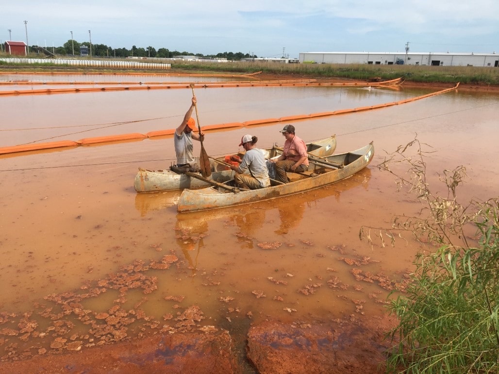 Three workers in a double canoe boat work to collect field samples in shallow orange water. The land is sectioned in long rows separated by orange floating dividers.