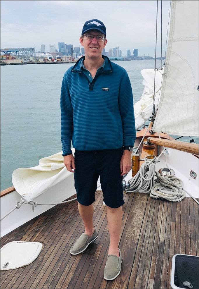 A man standing on the prow of a sailboat with Boston Harbor in the background. The man, wearing glasses, is dressed in a blue baseball cap, blue quarter zip long sleeved jacket, dark knee shorts and tan shoes.