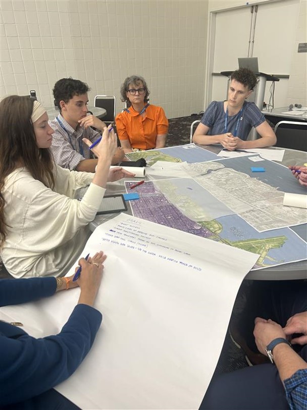 A number of workshop participants are seated around a table with materials on it; one taking notes on a large piece of paper.