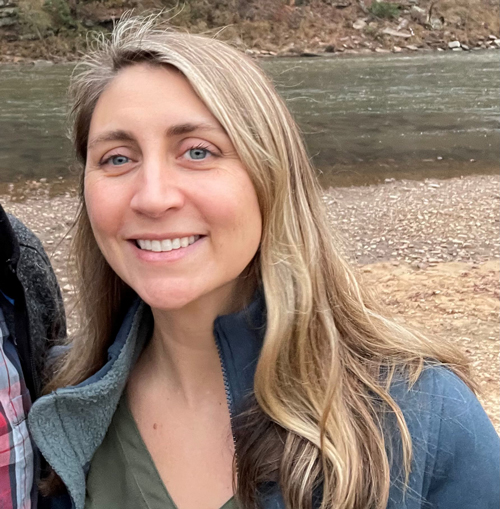 A blond woman with log hair and blue eyes smiling at the camera, wearing a blue jacket and green v-neck shirt. Gravel and a stream are in the background.