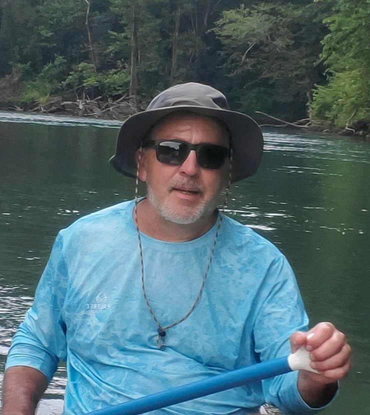 A man in a row boat holding an oar wearing a light blue shirt sunglasses and a grey floppy hat with cord on a body of green water. A tree line with woddy debris is in the background.
