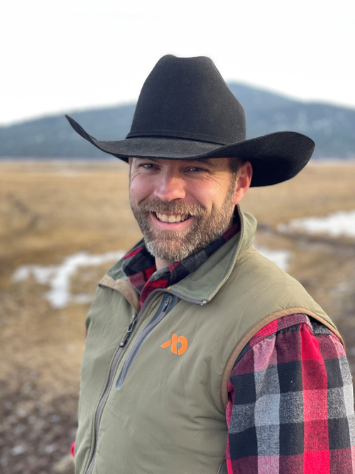 A smiling man wearing a dark grey cowbow style hat, red and black plaid shirt and tan vest stands. A blurred background contains some water or snow patches on a brown landscape and a mountain in the distance.