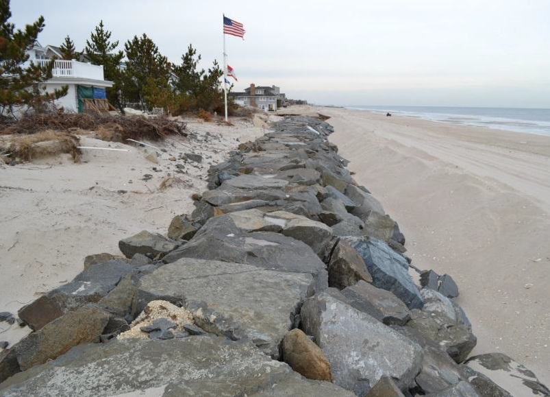 Exposed rock barrier away from shore. To the left is are homes and a flag pole; to the right is a stretch of sand beach and the water.