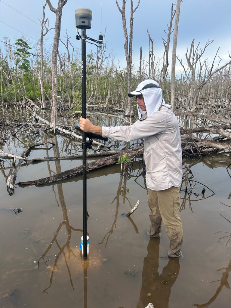 A person wearing a hat and long-sleeve shirt stands in shallow water among dead mangrove trees, holding and stabilizing a tall surveying instrument mounted on a pole.