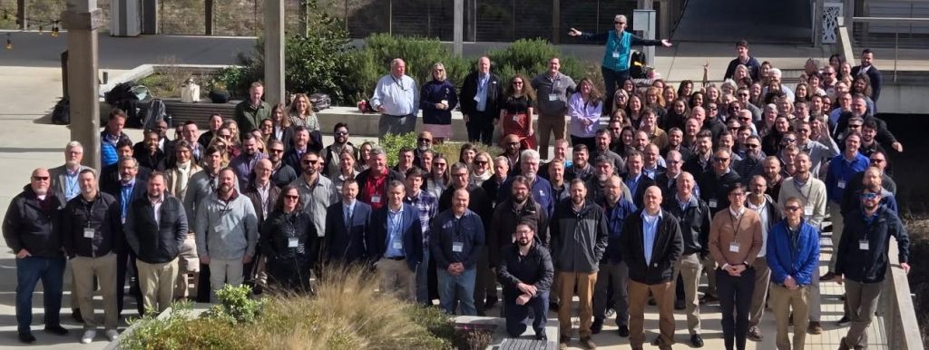A large group of attendees at the USACE South Atlantic Division (SAD) Coastal Summit in 2025, posing for a group photograph outdoors on a sunny day. The diverse group is arranged on steps in a modern plaza, smiling for the camera.