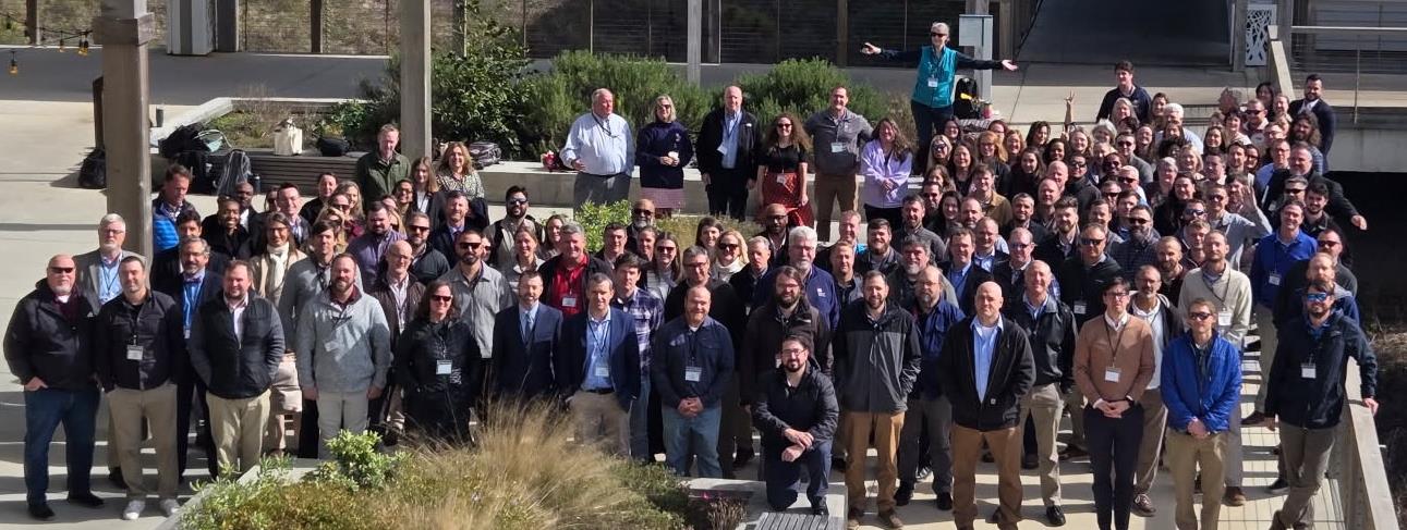 A large group of attendees at the USACE South Atlantic Division (SAD) Coastal Summit in 2025, posing for a group photograph outdoors on a sunny day. The diverse group is arranged on steps in a modern plaza, smiling for the camera.
