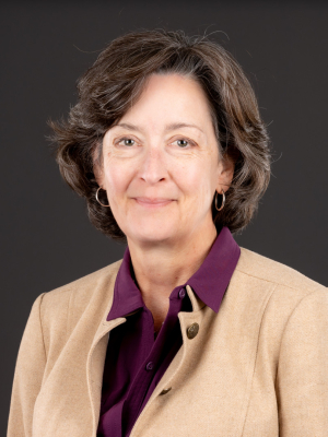 Professional head-and-shoulders portrait of a woman with short brown hair, wearing a tan blazer over a dark purple collared shirt against a dark background.